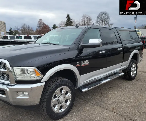 A front three-quarter view of a black and silver heavy-duty pickup truck with a matching camper shell, parked in a lot under a cloudy sky.